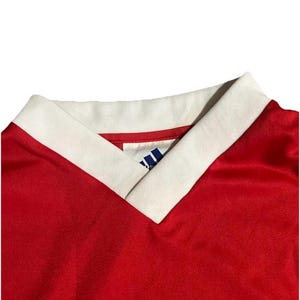 K&ouml;nnte beinhalten: Nahaufnahme eines roten Sporttrikots mit wei&szlig;em V-Ausschnitt. Der Kragen hat eine wei&szlig;e Einfassung. Das Adidas-Logo ist auf dem Trikot sichtbar. Das Trikot besteht aus einem gl&auml;nzenden Material.