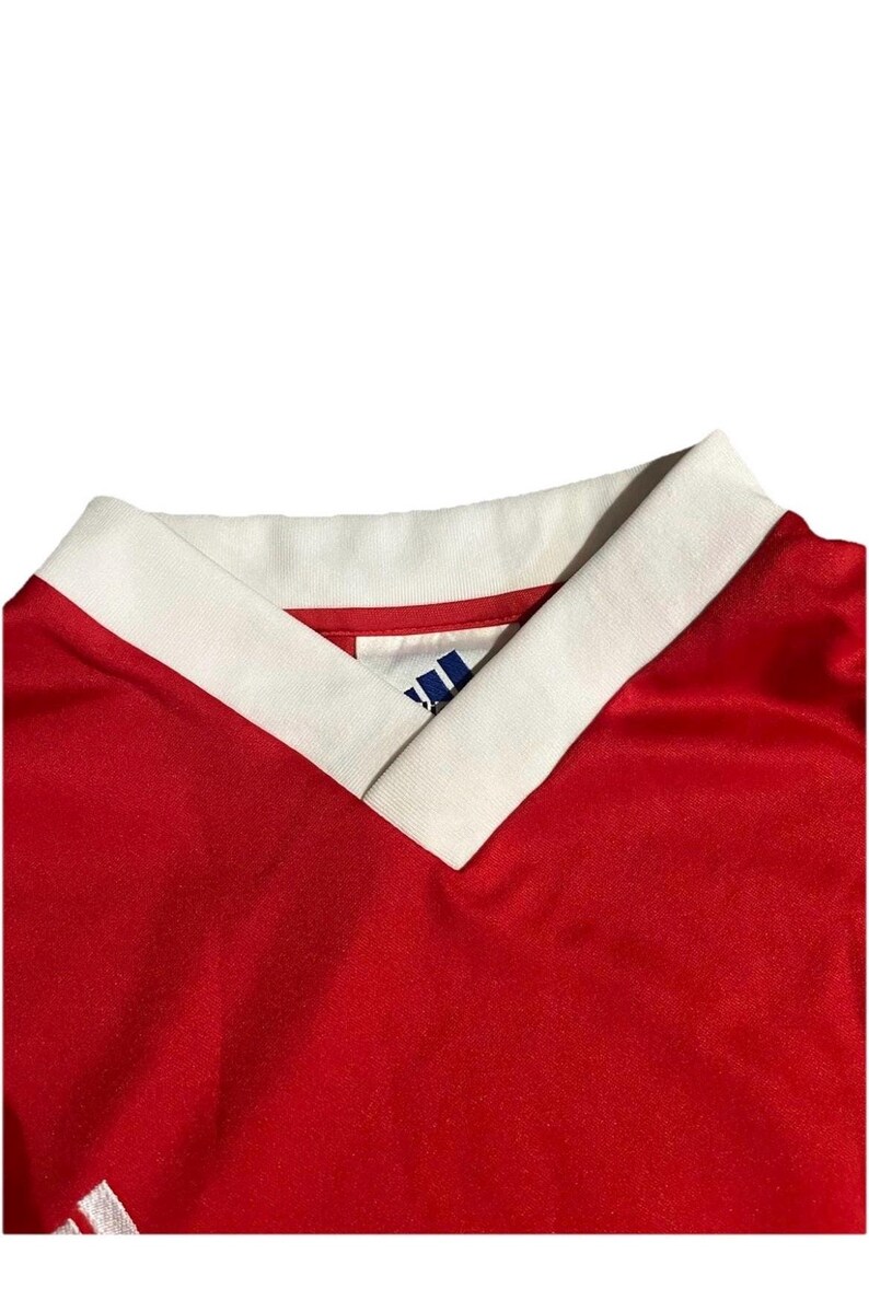 K&ouml;nnte beinhalten: Nahaufnahme eines roten Sporttrikots mit wei&szlig;em V-Ausschnitt. Der Kragen hat eine wei&szlig;e Einfassung. Das Adidas-Logo ist auf dem Trikot sichtbar. Das Trikot besteht aus einem gl&auml;nzenden Material.