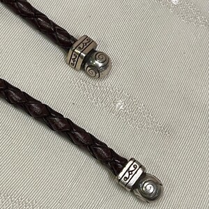 Może przedstawiać: Zbliżenie dw&oacute;ch ciemnobrązowych plecionych sk&oacute;rzanych sznurk&oacute;w ze srebrnymi metalowymi akcentami. Metalowe elementy mają ozdobny wz&oacute;r i spiralny detal. Sznurki są prawdopodobnie częścią naszyjnika lub bransoletki.