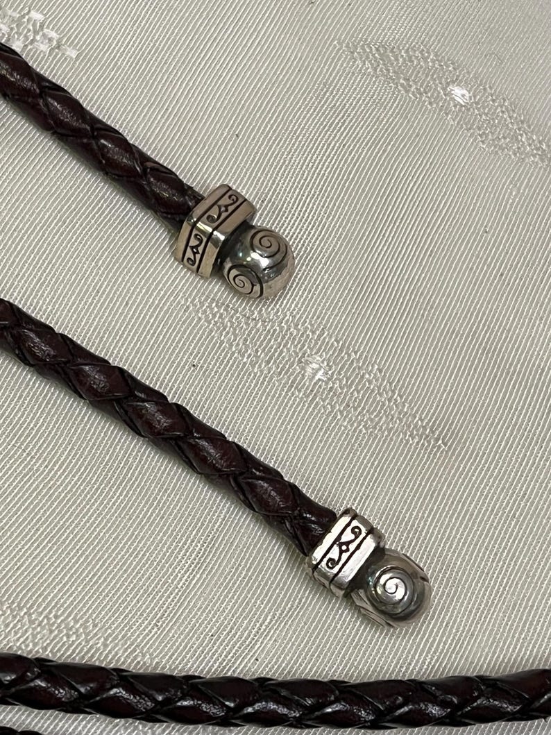 Może przedstawiać: Zbliżenie dw&oacute;ch ciemnobrązowych plecionych sk&oacute;rzanych sznurk&oacute;w ze srebrnymi metalowymi akcentami. Metalowe elementy mają ozdobny wz&oacute;r i spiralny detal. Sznurki są prawdopodobnie częścią naszyjnika lub bransoletki.