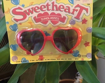 May include: Red heart-shaped sunglasses with a yellow and blue packaging that says "Sweetheart Sunglasses" and "For ages 3 & over".