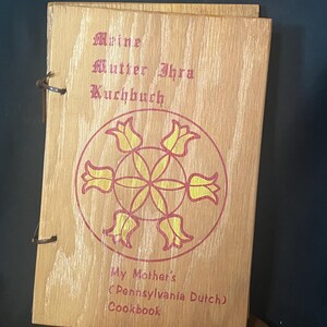 May include: A wooden cookbook with a light brown grain finish, bound by a dark leather cord. The cover features the text "Meine Mutter Ihra Kuchbuch" and "My Mother's (Pennsylvania Dutch) Cookbook" in red, with a central yellow and red floral design.