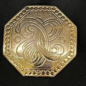 May include: Gold-toned, octagonal brooch with an ornate, swirling design in the center. The brooch is edged with a border of small, raised circles. The background is a dark, solid color.