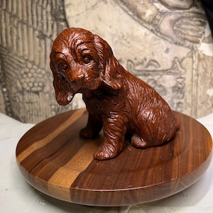 May include: A detailed, brown wooden sculpture of a dog sitting on a round, wooden base. The dog has long, floppy ears and a gentle expression. The base is made of alternating dark and light wood tones.