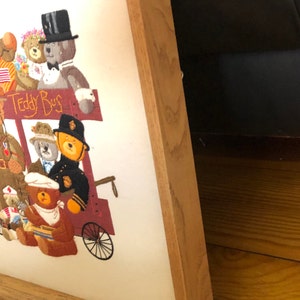 May include: A framed embroidery of a group of teddy bears riding a red wagon with the words "Teddy Bus" on the side. The embroidery is in a wooden frame with a light brown finish.
