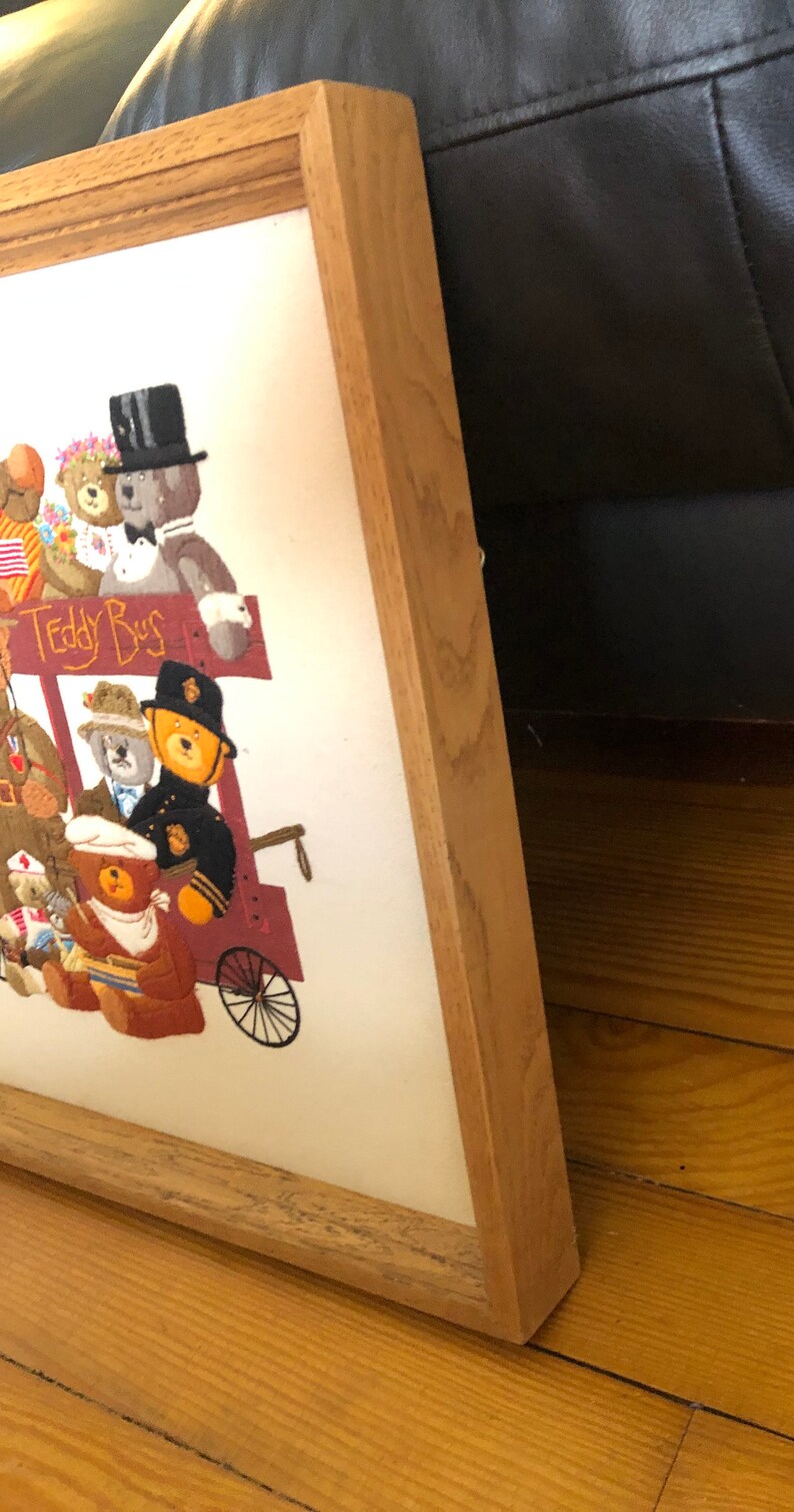 May include: A framed embroidery of a group of teddy bears riding a red wagon with the words "Teddy Bus" on the side. The embroidery is in a wooden frame with a light brown finish.