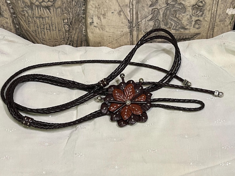 Może przedstawiać: Brązowy sk&oacute;rzany krawat bolo z ozdobną zawieszką w kształcie kwiatu. Zawieszka posiada warstwowe, liściaste elementy w odcieniach brązu i czerwieni, z małym, srebrnym klejnotem pośrodku. Pleciony sk&oacute;rzany sznurek ma srebrne końc&oacute;wki.