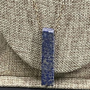 May include: A lapis lazuli pendant necklace. The rectangular pendant, in a deep blue hue with gold flecks, hangs from a fine gold-coloured chain. The necklace is presented against a textured, neutral-toned background.