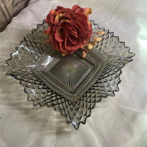 May include: A gray, diamond-shaped glass dish with a textured surface and a decorative edge. A large, red and yellow artificial rose rests in the dish. The dish is on a white, textured surface.