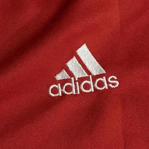 K&ouml;nnte beinhalten: Nahaufnahme eines roten Stoffes mit dem wei&szlig;en Adidas-Logo, das oben links eingestickt ist. Das Logo zeigt drei schr&auml;ge Streifen &uuml;ber dem Wort "adidas" in einer serifenlosen Schrift.