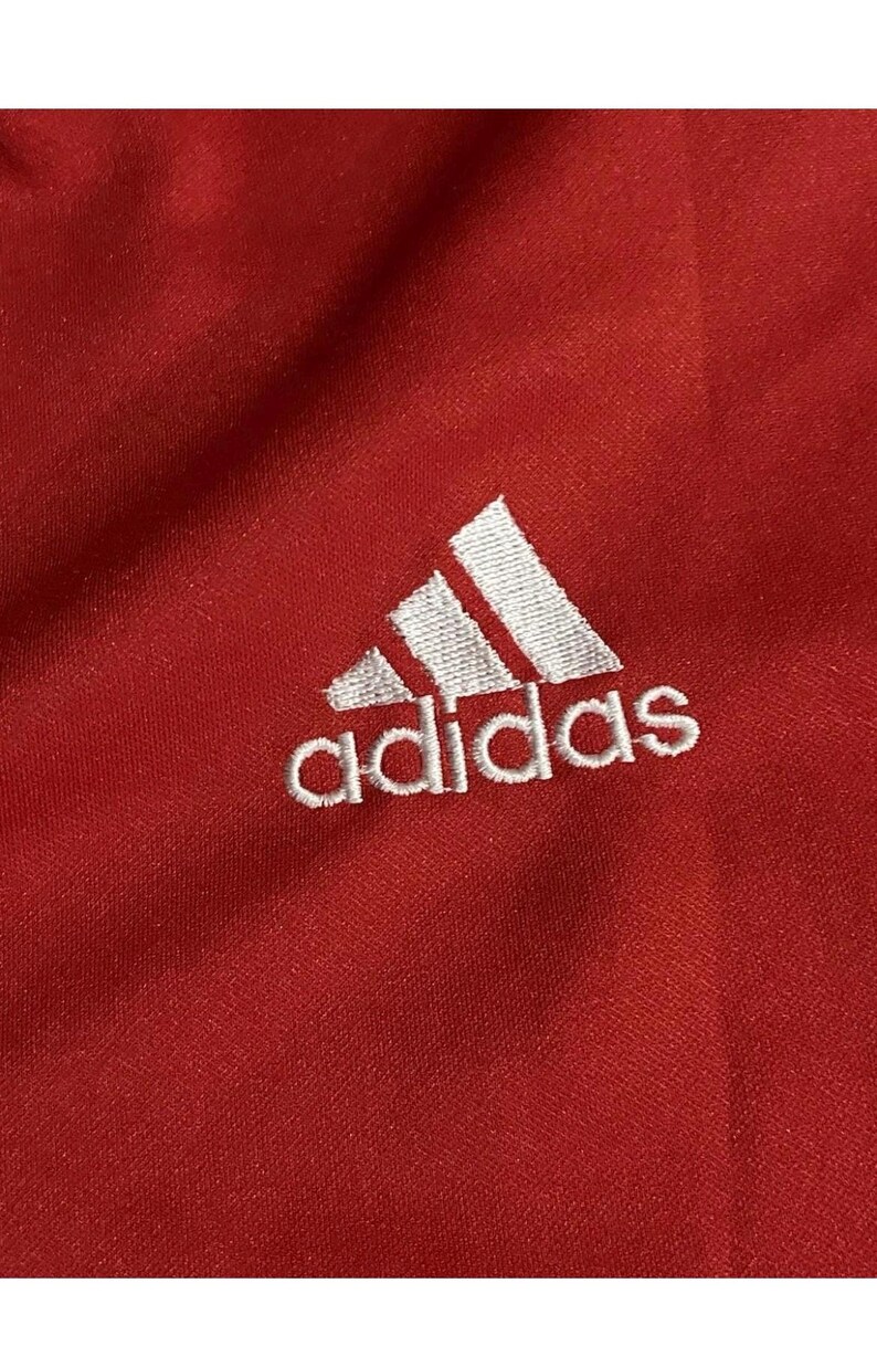 K&ouml;nnte beinhalten: Nahaufnahme eines roten Stoffes mit dem wei&szlig;en Adidas-Logo, das oben links eingestickt ist. Das Logo zeigt drei schr&auml;ge Streifen &uuml;ber dem Wort "adidas" in einer serifenlosen Schrift.