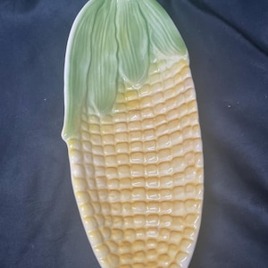 May include: A ceramic serving dish shaped like an ear of corn. The dish features a textured surface with yellow kernels and light green husks at the top. The elongated, oval-shaped piece has a glossy finish and mimics the appearance of corn.