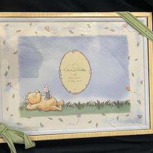 Könnte beinhalten: Ein rechteckiger Bilderrahmen mit einer Winnie Puuh und Ferkel Illustration auf hellblauem und cremefarbenem Hintergrund. Die Mitte enthält einen ovalen Rahmen mit dem Text 'a place photo of baby here'. Eine hellgrüne Schleife ist an der Ecke befestigt.