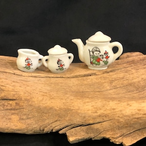 May include: A miniature white porcelain tea set with a teapot, creamer, and sugar bowl. Each piece features a hand-painted design of two children in green and red clothing.