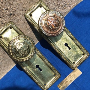 Könnte beinhalten: Zwei antike Türklinken aus Messing mit Schlüsselloch und verzierten Details. Die Knöpfe zeigen ein erhabenes Monogramm mit den Buchstaben "HS". Eine Messingplakette trägt die Aufschrift "HIGH SCHOOL 1884-1896 READING, PA".
