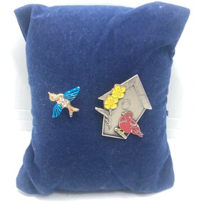 Puede incluir: Dos pines de esmalte sobre un fondo de terciopelo azul. Un pin presenta un pájaro azul con un cuerpo de pedrería y el otro pin presenta un pájaro rojo posado en una rama frente a una casita de pájaros amarilla con flores amarillas.