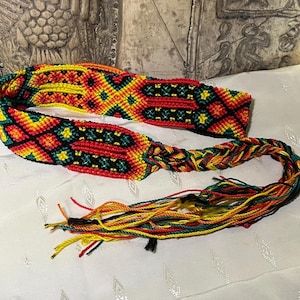 May include: A colorful woven belt with a geometric pattern in red, yellow, green, and black. The belt has long tassels in the same colors.