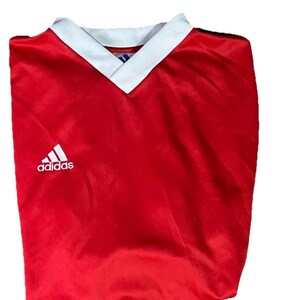 K&ouml;nnte beinhalten: Ein rotes Adidas Fu&szlig;balltrikot mit wei&szlig;em V-Ausschnitt und Adidas-Logo. Das Trikot ist gefaltet und scheint aus einem glatten, m&ouml;glicherweise synthetischen Stoff zu sein.