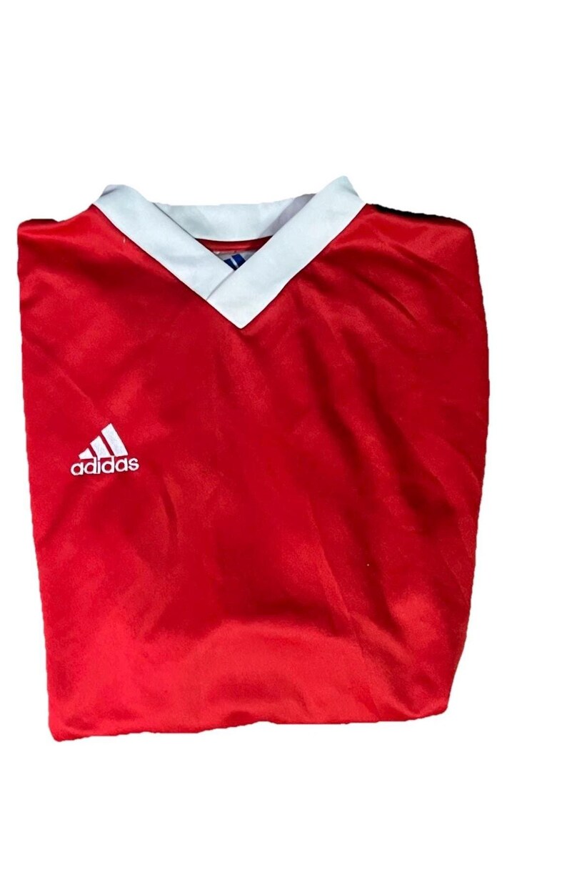 K&ouml;nnte beinhalten: Ein rotes Adidas Fu&szlig;balltrikot mit wei&szlig;em V-Ausschnitt und Adidas-Logo. Das Trikot ist gefaltet und scheint aus einem glatten, m&ouml;glicherweise synthetischen Stoff zu sein.