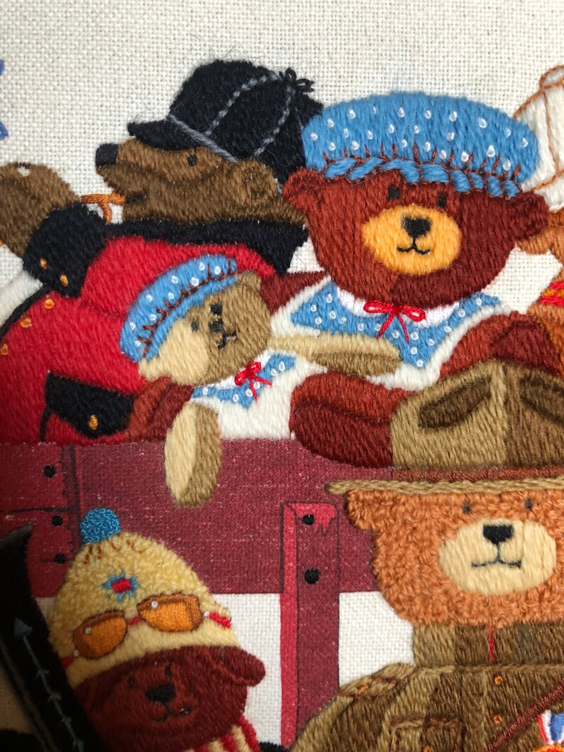 May include: Embroidered wall art featuring a group of teddy bears wearing different hats and outfits. The bears are arranged in a row, with the one in the front wearing a blue hat and a red bow tie. The bears are all smiling and looking at the viewer.