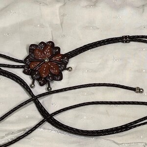 Może przedstawiać: Krawat bolo z wisiorkiem w kształcie kwiatu z brązowymi i bordowymi sk&oacute;rzanymi płatkami. Wisiorek ma srebrny środek i jest przymocowany do czarnego plecionego sznurka ze srebrnymi końc&oacute;wkami. Całość na białym tle.