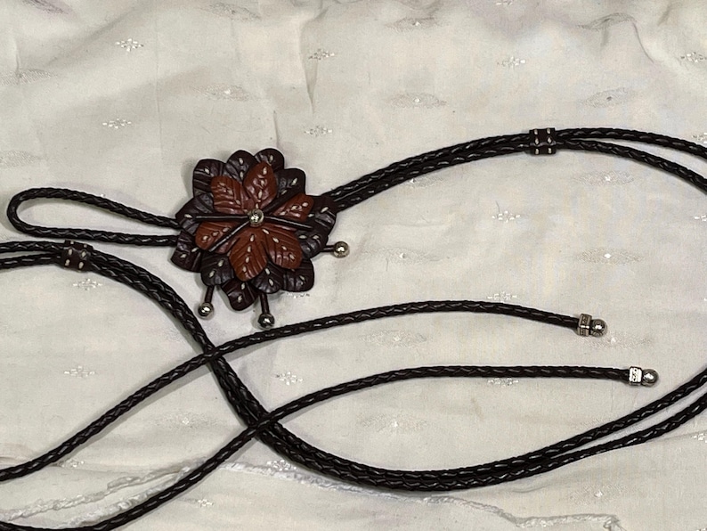 Może przedstawiać: Krawat bolo z wisiorkiem w kształcie kwiatu z brązowymi i bordowymi sk&oacute;rzanymi płatkami. Wisiorek ma srebrny środek i jest przymocowany do czarnego plecionego sznurka ze srebrnymi końc&oacute;wkami. Całość na białym tle.