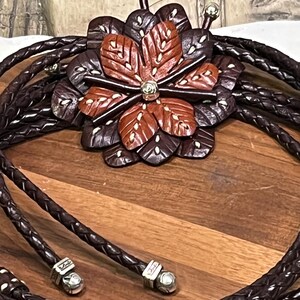 Może przedstawiać: Brązowy sk&oacute;rzany krawat bolo z wisiorkiem w kształcie kwiatu. Wisiorek ma centralny kwiat w odcieniach brązu i czerwieni z białymi akcentami, otoczony ciemniejszymi brązowymi płatkami. Pleciony sk&oacute;rzany sznurek zakończony srebrnymi metalowymi końc&oacute;wkami.