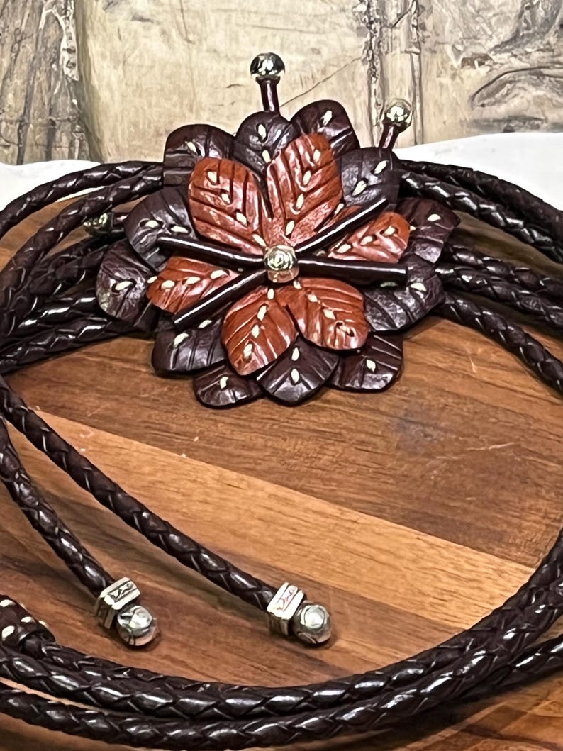 Może przedstawiać: Brązowy sk&oacute;rzany krawat bolo z wisiorkiem w kształcie kwiatu. Wisiorek ma centralny kwiat w odcieniach brązu i czerwieni z białymi akcentami, otoczony ciemniejszymi brązowymi płatkami. Pleciony sk&oacute;rzany sznurek zakończony srebrnymi metalowymi końc&oacute;wkami.