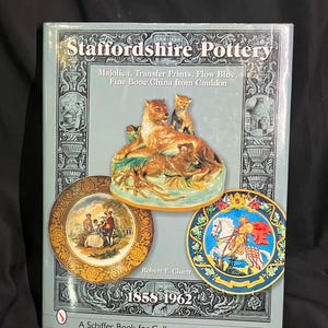 Puede incluir: Un libro titulado "Staffordshire Pottery" con una cubierta azul claro. La portada presenta imágenes de cerámica, incluyendo una figura de león y cachorro, y platos decorativos. El texto incluye "Majolica, Transfer Prints, Flow Blue" y "1858-1962".