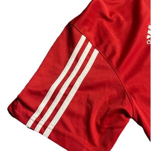 K&ouml;nnte beinhalten: Roter Sport&auml;rmel mit drei wei&szlig;en Streifen. Der &Auml;rmel besteht aus einem gl&auml;nzenden, strukturierten Material. Das Adidas-Logo ist in der oberen rechten Ecke sichtbar.