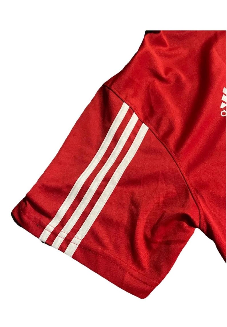 K&ouml;nnte beinhalten: Roter Sport&auml;rmel mit drei wei&szlig;en Streifen. Der &Auml;rmel besteht aus einem gl&auml;nzenden, strukturierten Material. Das Adidas-Logo ist in der oberen rechten Ecke sichtbar.