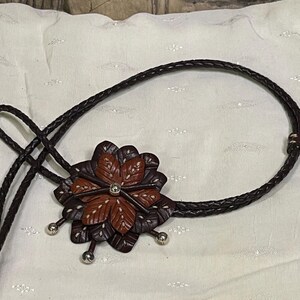 Może przedstawiać: Brązowy sk&oacute;rzany krawat bolo z wisiorkiem w kształcie kwiatu. Wisiorek ma ciemnobrązowe i czerwonobrązowe płatki z białymi przeszyciami i centralnym klejnotem. Pleciony sznurek jest ciemnobrązowy ze srebrnymi akcentami.