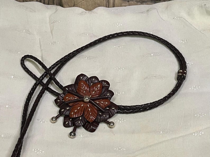 Może przedstawiać: Brązowy sk&oacute;rzany krawat bolo z wisiorkiem w kształcie kwiatu. Wisiorek ma ciemnobrązowe i czerwonobrązowe płatki z białymi przeszyciami i centralnym klejnotem. Pleciony sznurek jest ciemnobrązowy ze srebrnymi akcentami.