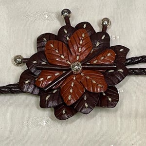 Może przedstawiać: Brązowy sk&oacute;rzany krawat bolo z kwiatowym wzorem. Kwiat ma ciemnobrązowe i czerwonobrązowe płatki z białymi przeszyciami. Srebrne metalowe akcenty znajdują się na środku kwiatu i końcach krawata. Pleciony sk&oacute;rzany sznurek dopełnia projekt.