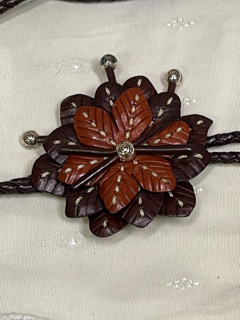 Może przedstawiać: Brązowy sk&oacute;rzany krawat bolo z kwiatowym wzorem. Kwiat ma ciemnobrązowe i czerwonobrązowe płatki z białymi przeszyciami. Srebrne metalowe akcenty znajdują się na środku kwiatu i końcach krawata. Pleciony sk&oacute;rzany sznurek dopełnia projekt.