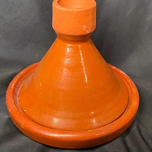 May include: An orange-colored tagine pot with a conical lid and a wide, shallow base. The lid has a cylindrical handle at the top. The pot is made of terracotta and has a smooth, glazed finish. The tagine is designed for slow cooking.
