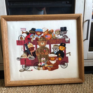 May include: A framed needlepoint embroidery depicting a red and white bus filled with teddy bears. The bus has the words "Teddy Bus" on the side. The teddy bears are wearing various outfits, including a military uniform, a sailor outfit, and a chef's hat.