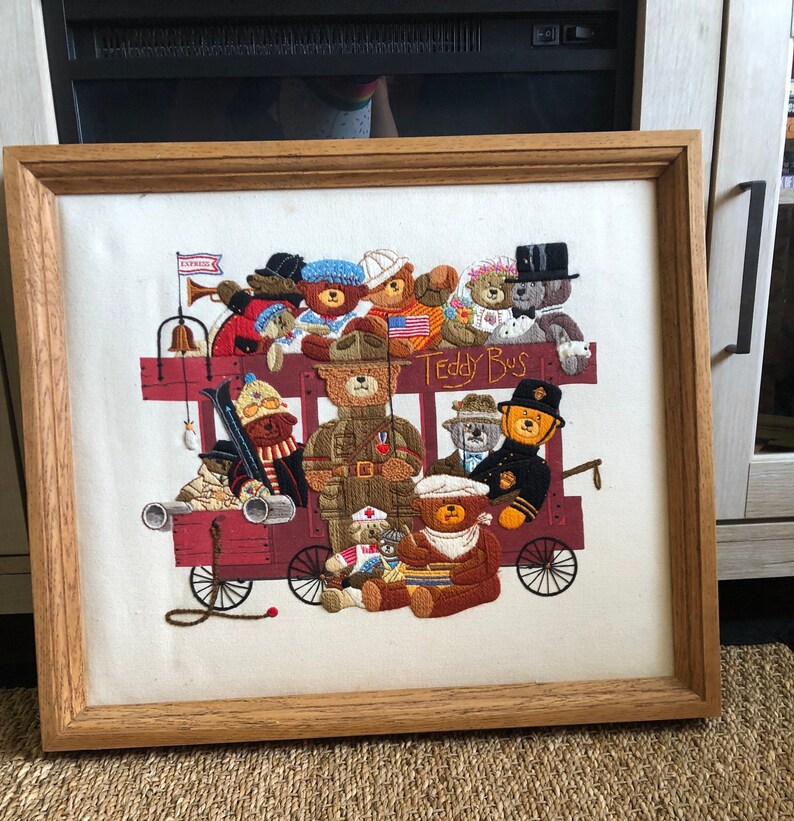 May include: A framed needlepoint embroidery depicting a red and white bus filled with teddy bears. The bus has the words "Teddy Bus" on the side. The teddy bears are wearing various outfits, including a military uniform, a sailor outfit, and a chef's hat.