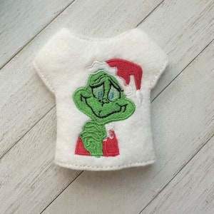 May include: White felt shirt with an embroidered image of the Grinch wearing a Santa hat. The Grinch is green with a red and white Santa hat.