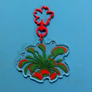 May include: A clear acrylic keychain featuring a printed illustration of a green Venus flytrap plant with red-rimmed, toothy traps. The keychain is attached to a bright red, flower-shaped plastic clasp and a circular jump ring connector.
