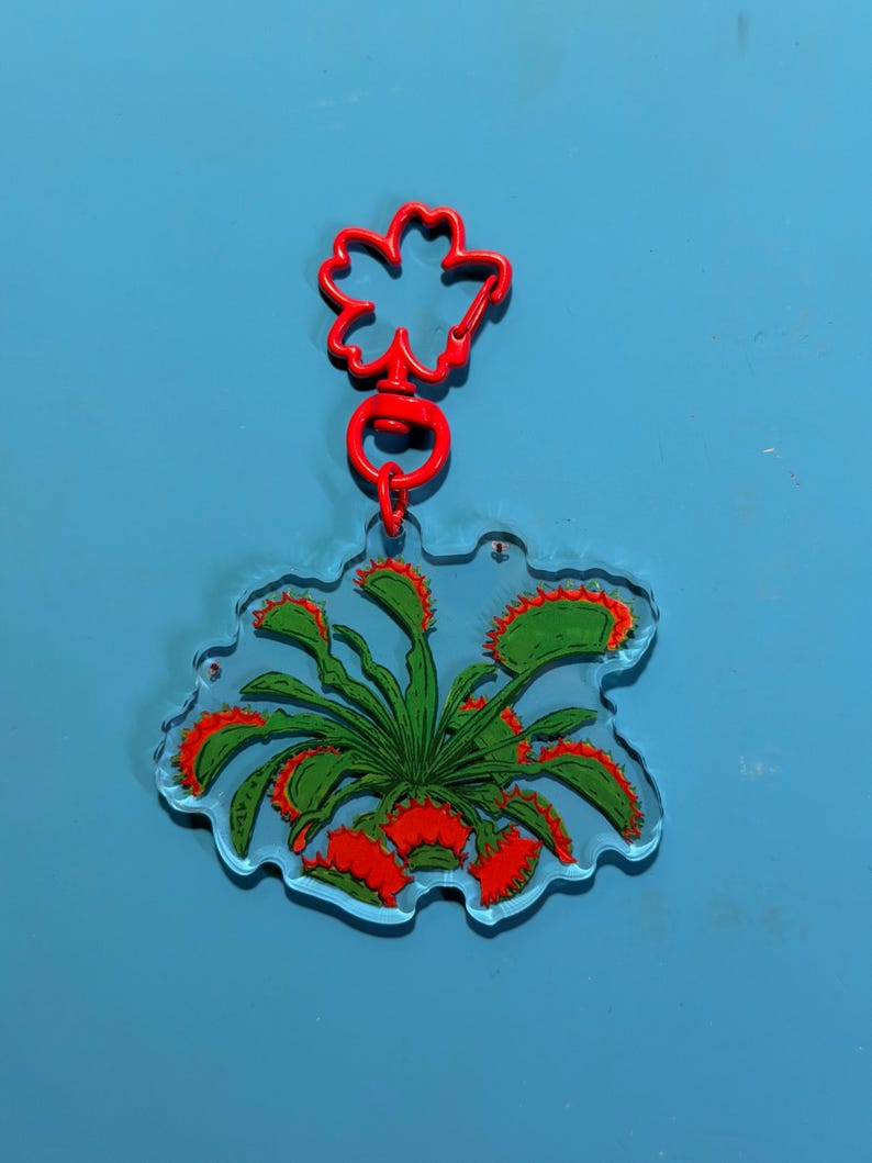 May include: A clear acrylic keychain featuring a printed illustration of a green Venus flytrap plant with red-rimmed, toothy traps. The keychain is attached to a bright red, flower-shaped plastic clasp and a circular jump ring connector.