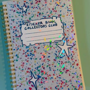 May include: A spiral-bound notebook with a light blue cover featuring a multicolored confetti pattern and holographic accents. The cover displays a rectangular label with the text "STICKER BOOK COLLECTORS CLUB" and three hand-drawn star shapes.