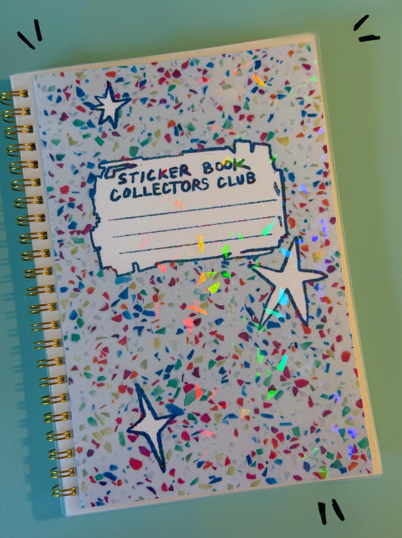 May include: A spiral-bound notebook with a light blue cover featuring a multicolored confetti pattern and holographic accents. The cover displays a rectangular label with the text "STICKER BOOK COLLECTORS CLUB" and three hand-drawn star shapes.
