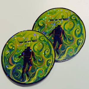 Puede incluir: Dos pegatinas circulares con un diseño vibrante. Cada una presenta un astronauta silueteado sobre un fondo arremolinado verde y amarillo con el texto "I'll do my best". Los acentos holográficos añaden brillo.