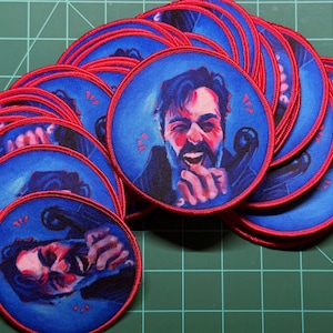 May include: A collection of circular embroidered patches featuring a stylized illustration of a person with a beard laughing while holding a telephone receiver. The patches have a vibrant blue background, red accents, and a stitched red border.