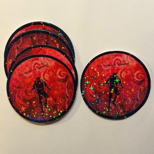 May include: A stack of round stickers with a red background and a swirling pattern. Each sticker features a black astronaut figure and the text "I'M HAVING A MOMENT." Holographic stars and accents add sparkle.