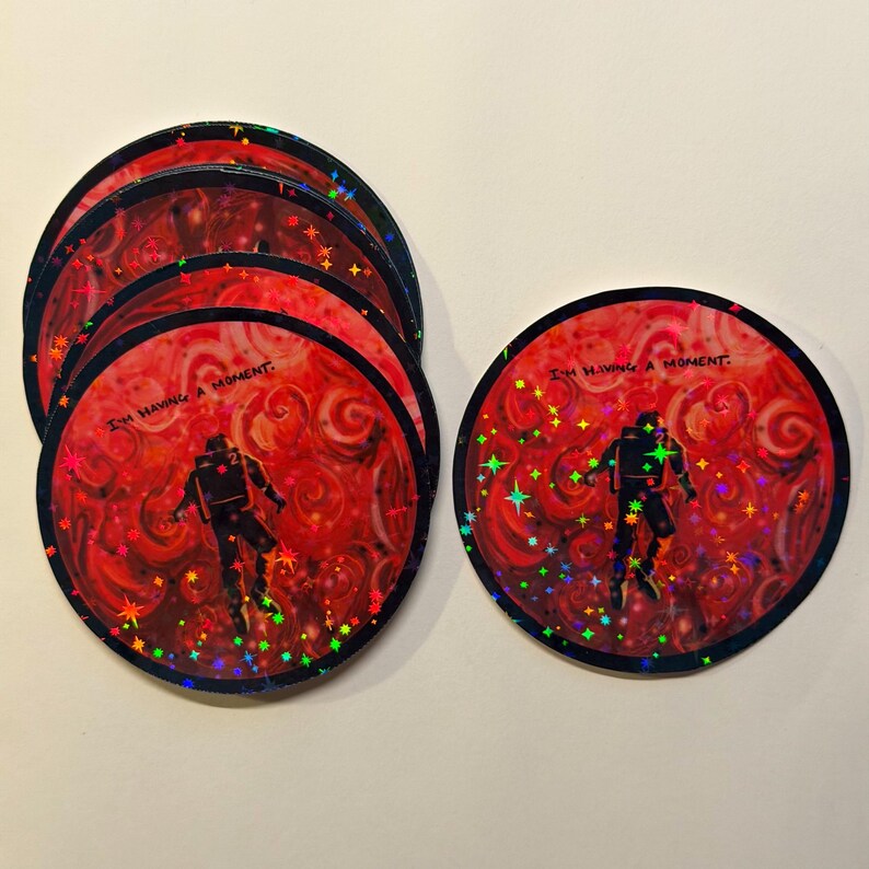 May include: A stack of round stickers with a red background and a swirling pattern. Each sticker features a black astronaut figure and the text "I'M HAVING A MOMENT." Holographic stars and accents add sparkle.