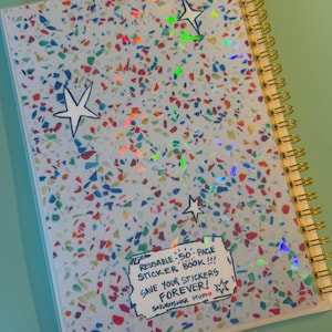 May include: A spiral-bound sticker book with a light blue cover featuring a multicolored confetti pattern and holographic star accents. A white rectangular label on the cover reads: REUSABLE 50-PAGE STICKER BOOK!!! SAVE YOUR STICKERS FOREVER! SATURNSWAR STUDIO.