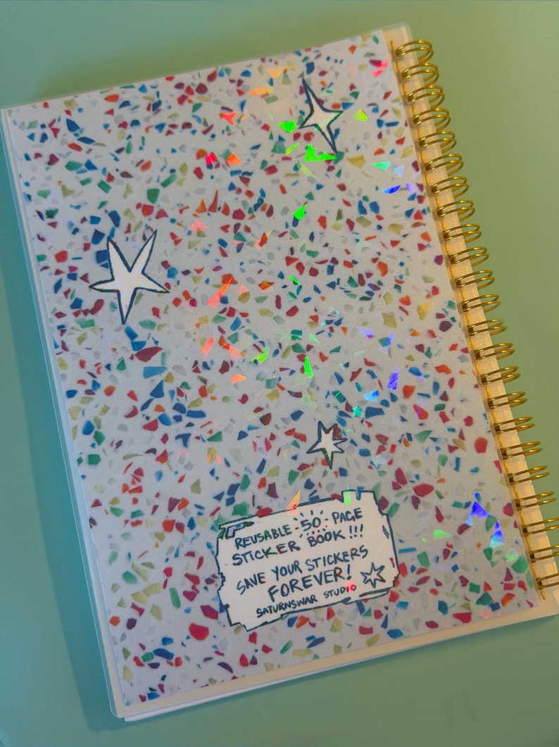 May include: A spiral-bound sticker book with a light blue cover featuring a multicolored confetti pattern and holographic star accents. A white rectangular label on the cover reads: REUSABLE 50-PAGE STICKER BOOK!!! SAVE YOUR STICKERS FOREVER! SATURNSWAR STUDIO.