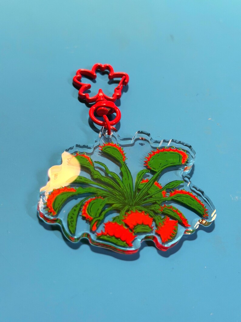 May include: A clear acrylic keychain featuring a printed illustration of a green Venus flytrap plant with vibrant red interior traps. The keychain is attached to a bright red, flower-shaped plastic clasp, providing a secure way to hang the item.